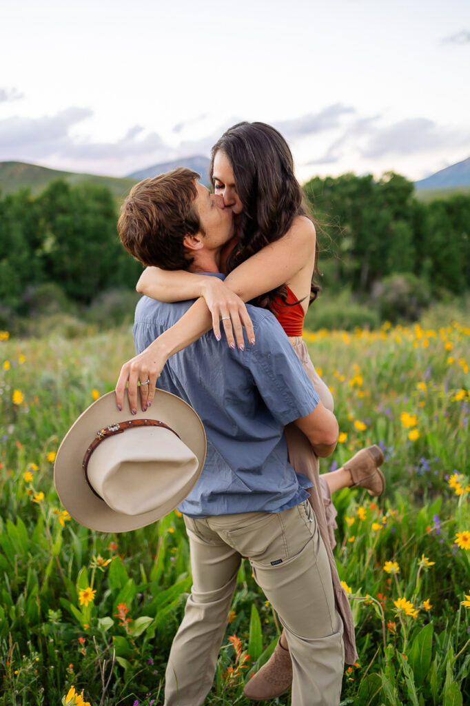 kissing couple in flower fields wildflowers holding hat About Us Bio Lydia + T anniversary - photo by Mountain Magic Media - Colorado Photographer Team Travel Photographers