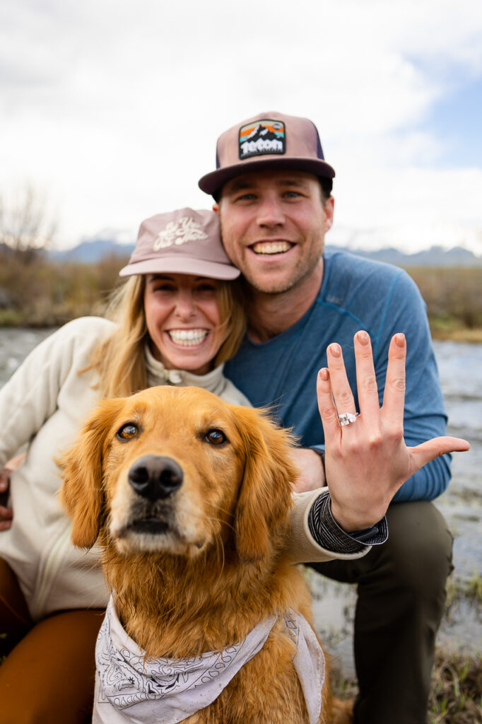 couple smiling holding up engagement ring with river stream in background and cute dog puppy face mountains with dog Crested Butte photographer Gunnison photographers Colorado photography - proposal engagement elopement wedding venue - photo by Mountain Magic Media