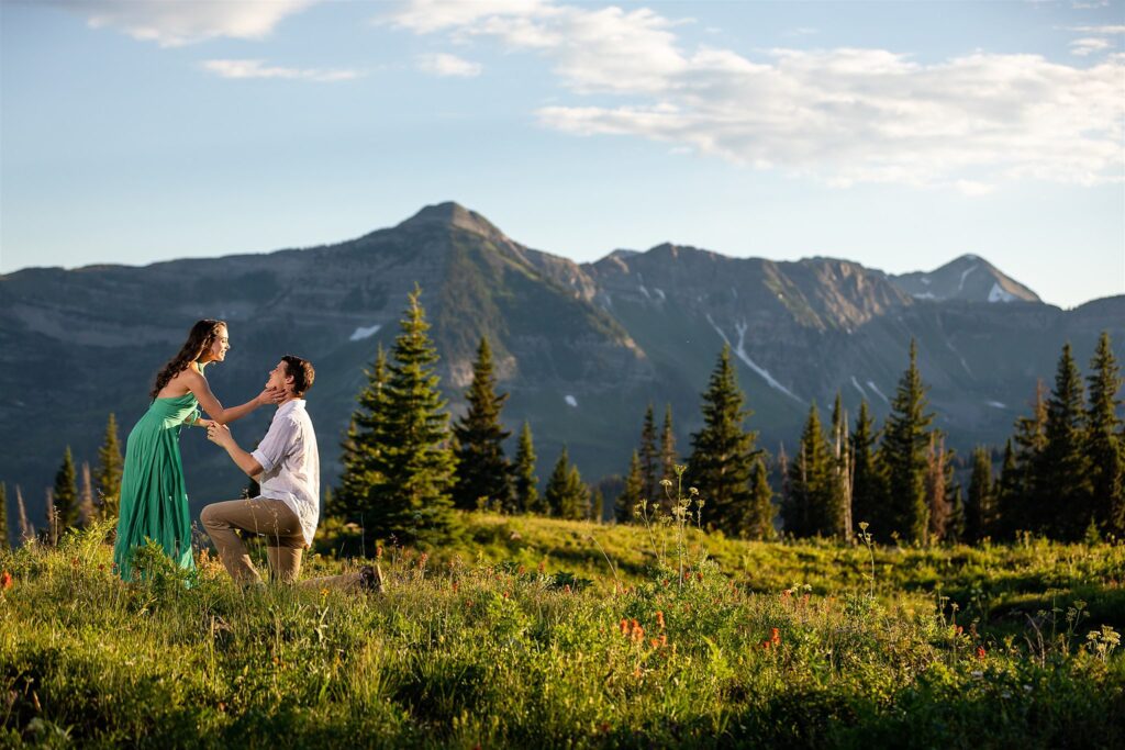 A woman in a green dress touches the face of her kneeling boyfriend in a remote mountain trail