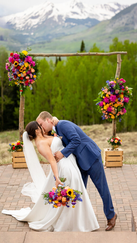 Bride and Groom married couple kissing under a wedding flowers arch with colorful floral bridal bouquet in Crested Butte at Ten Peaks wedding venue Umbrella Bar site.