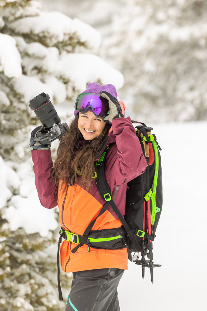 Woman smiling holding camera, one hand touching ski goggles, wearing backpack and bright orange and maroon coat in the snow.