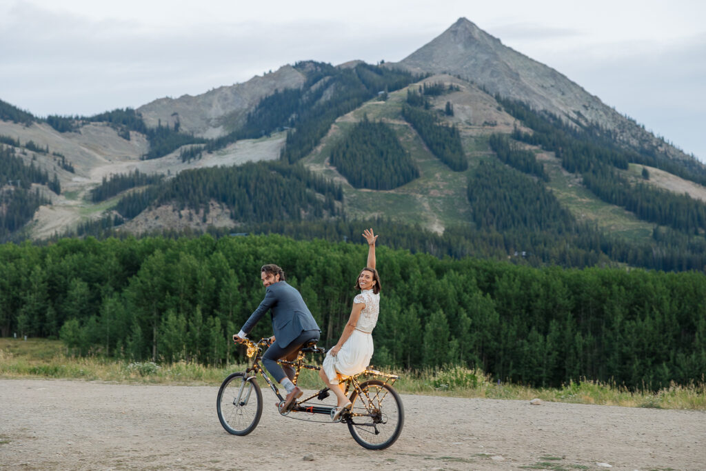 Newlweds Couple riding tandem bicycle with Mt. CB peak in the background at the Ten Peaks Umbrella Bar Wedding Venue in Crested Butte