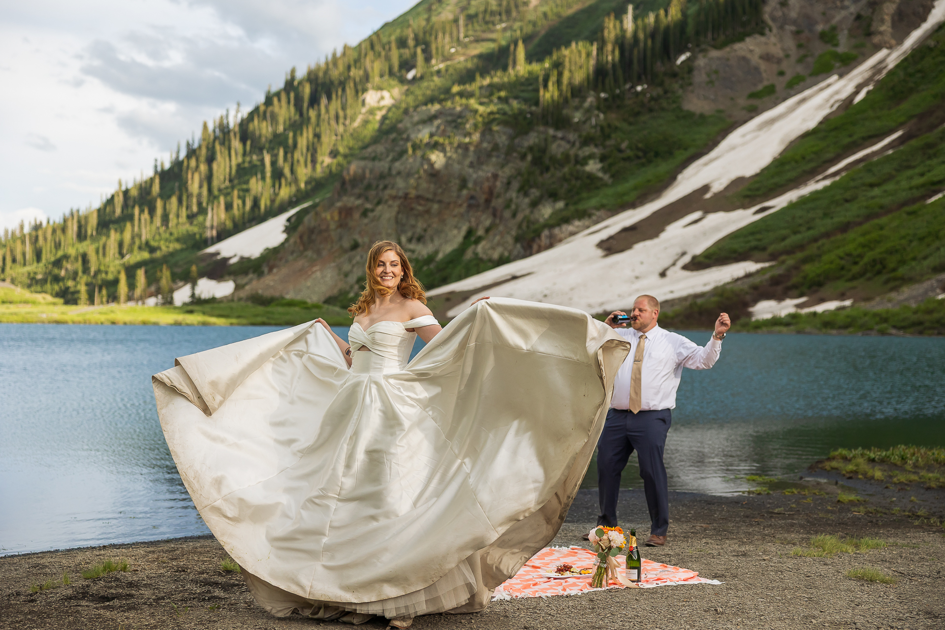 bride holding wedding dress while dancing at Emerald Lake picnic as groom celebrates in Crested Butte, Colorado