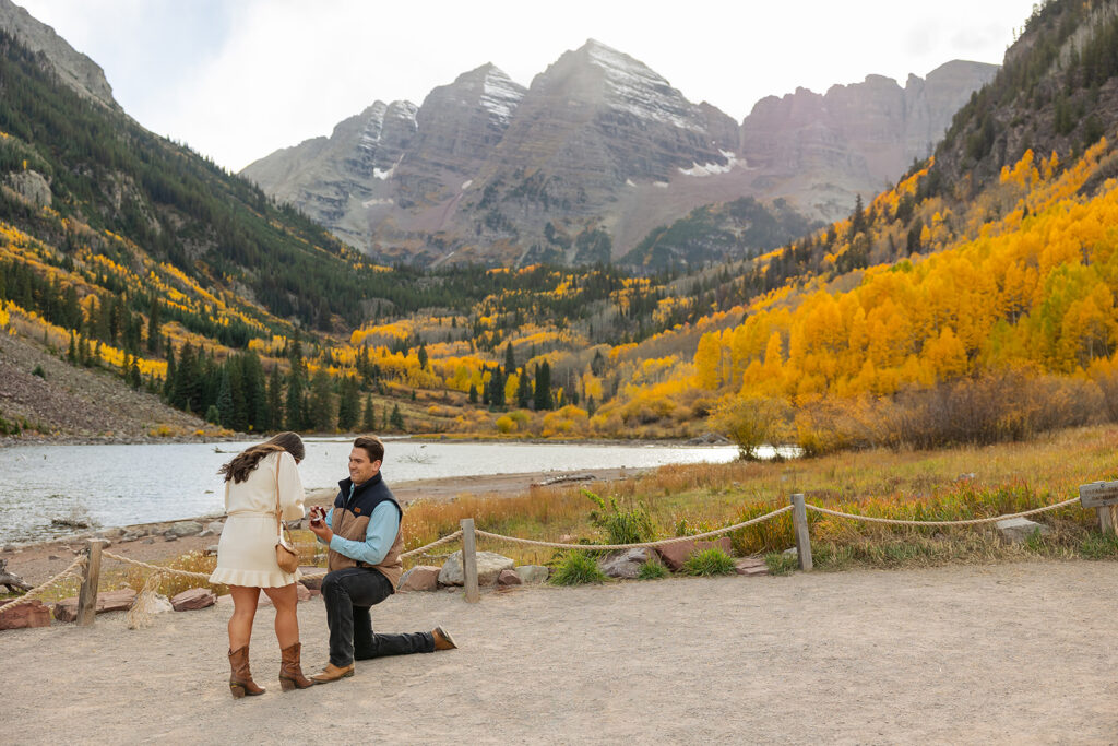 Surprise proposal at the Maroon Bells in Aspen Colorado — man on one knee at Maroon Lake during peak fall foliage, golden aspens and snow-capped Maroon Bells peaks behind them