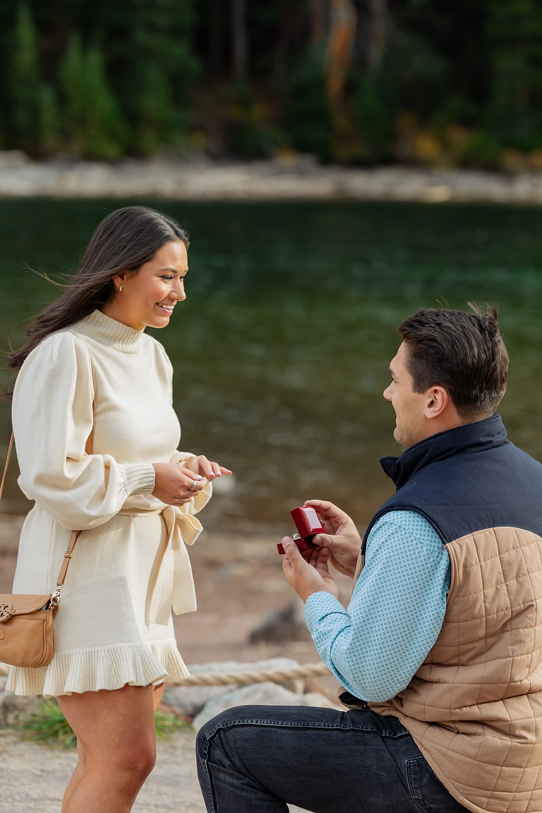 Close-up of a Maroon Bells proposal — man on one knee holding an open ring box as his partner smiles down at him, Maroon Lake and pine forest in the background