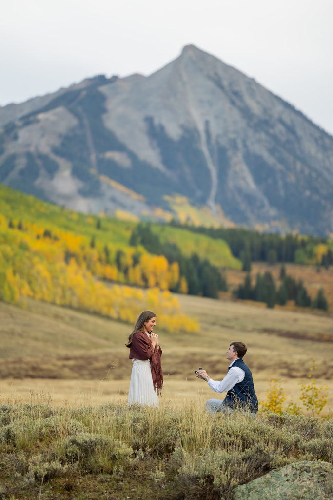 Man on one knee proposing to woman in a golden meadow below Mt. Crested Butte during peak fall foliage in Colorado man proposing to woman after a hike in Crested Butte surrounded by wildflowers with custom sign and florals for Proposal Packages