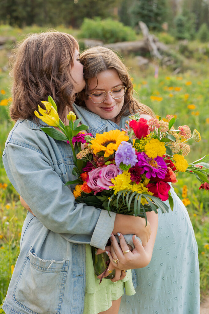 One woman kissing her newly engaged partner on the temple while she holds a large colorful bouquet of sunflowers, roses, and wildflowers in a Crested Butte summer meadow