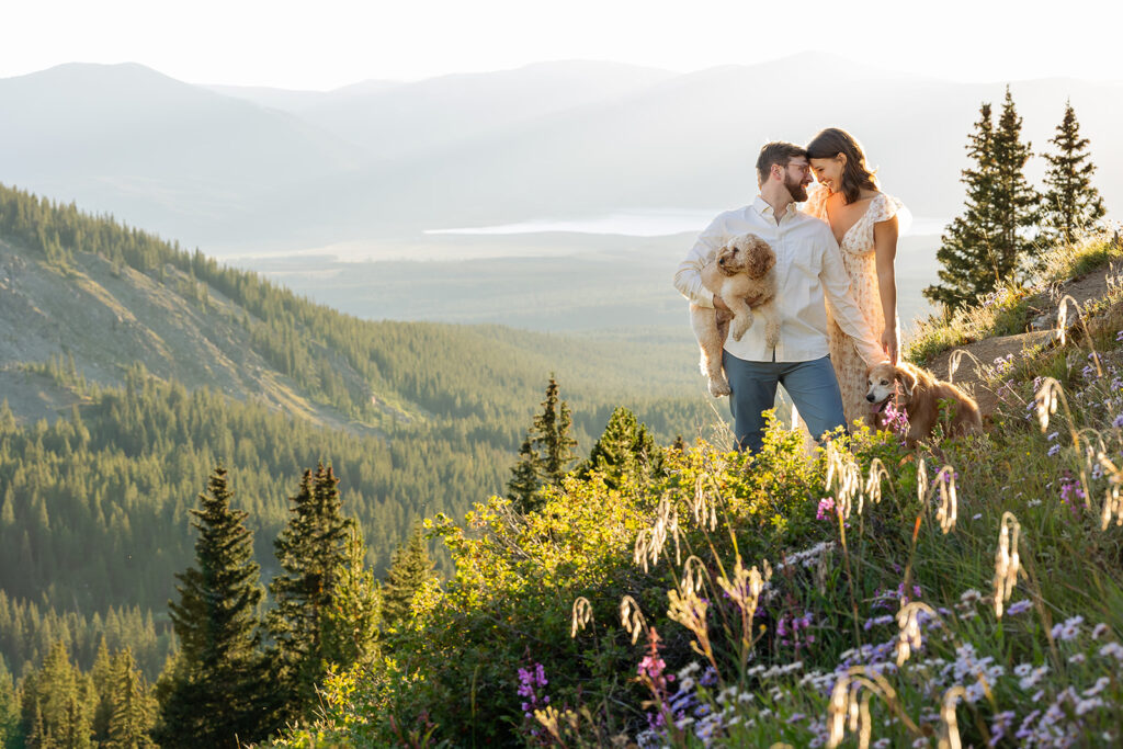 Engaged couple standing close and smiling at each other on a wildflower-covered hillside at Cottonwood Pass during sunrise, one partner holding a small dog while another dog stands nearby, sweeping valley and mountain views behind them, Crested Butte Colorado photographed by Mountain Magic Media