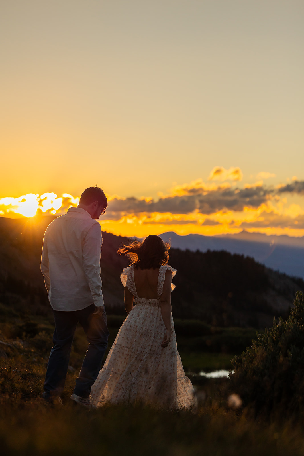 Engaged couple walking away from camera through alpine tundra at Cottonwood Pass during golden hour sunset, woman in a floral backless dress with hair blowing in the wind, small alpine lake visible below, Crested Butte Colorado