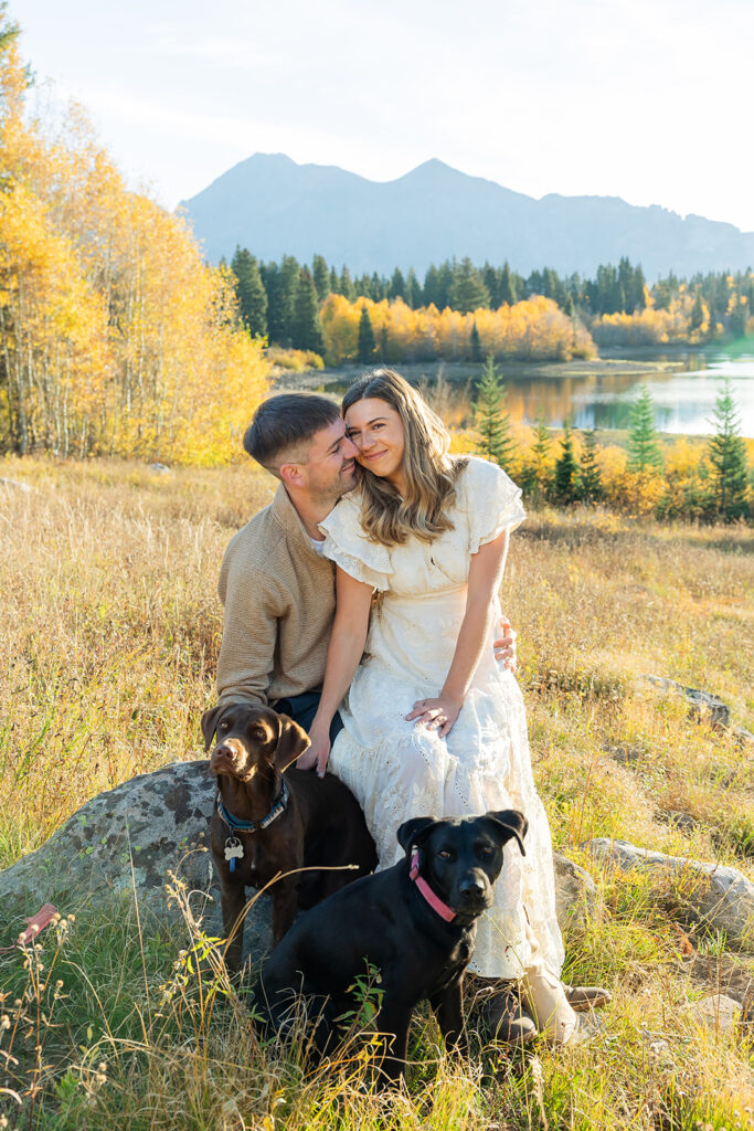Engaged couple seated together on a boulder in a golden meadow with their two dogs during a Kebler Pass fall engagement session, fall aspens and a mountain lake visible in the background, Crested Butte Colorado photographed by Mountain Magic Media