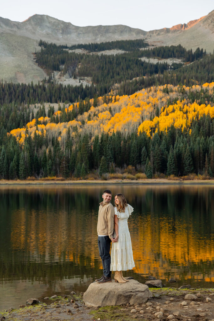Engaged couple standing on a boulder at Lost Lake Slough during a Kebler Pass fall engagement session, golden aspen trees reflected in still water with alpenglow on the mountains behind them, photographed by Mountain Magic Media Crested Butte Colorado