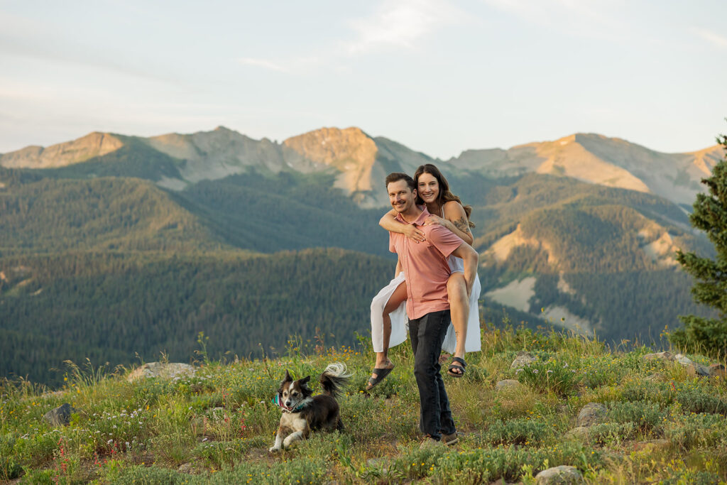 Engaged couple doing a piggyback pose on a wildflower ridge with their dog running beside them and a panoramic view of the Elk Mountains near Crested Butte Colorado